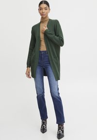 Cardigan verde realizzato in tessuto knit, design con apertura frontale, dotato di due tasche anteriori. Abbinato a jeans di denim blu e stivali neri alla caviglia.