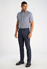 Camicia a maniche corte con bottoni e motivo a quadretti blu e bianchi, abbinata a pantaloni scuri su misura e mocassini neri.