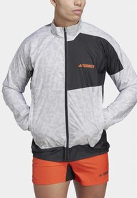 Leichte Jacke mit grauen und schwarzen Paneelen, horizontalem Streifenmuster, durchgehendem Reißverschluss und orangefarbenem "TERREX"-Logo auf der Brust.
