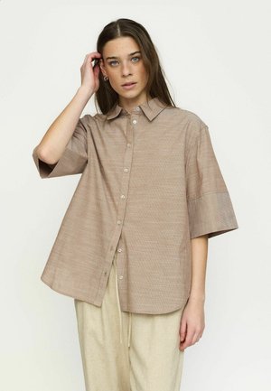 Young woman with long brown hair wearing a loose beige checkered short-sleeve button-up shirt and light-colored pants, standing against white background.