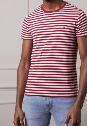 Man draagt een rood-wit horizontaal gestreept T-shirt met korte mouwen, gecombineerd met lichtblauwe spijkerbroek, staand binnenshuis tegen een neutrale achtergrond.