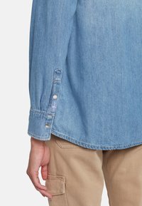 Camicia di denim azzurro chiaro con polsini abbottonati e orlo curvo, indossata sopra pantaloni cargo beige. Il tessuto ha una finitura morbida e texturizzata.