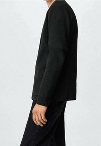 Blazer nero realizzato in tessuto testurizzato, con una vestibilità rilassata e tasche, indossato sopra pantaloni scuri, visto di lato.