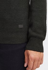 Maglione lavorato a maglia verde scuro con un motivo testurizzato, polsini a coste e una piccola etichetta nera con "ISould" ricamata di lato.