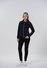 Veste matelassée bleu marine avec un col haut, fermeture éclair sur le devant, et accents blancs. Assortie à un pantalon slim noir et des baskets blanches.