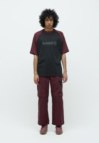 Young person with curly hair stands facing forward wearing a black and burgundy Timberland t-shirt, burgundy cargo pants, and matching shoes.