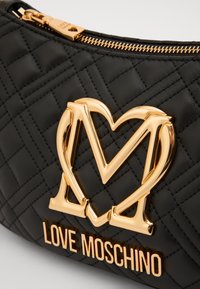 Crna jastučićasta sintetička torbica s istaknutim zlatnim srcem u obliku slova "M" i natpisom "LOVE MOSCHINO" u zlatnim metalik slovima.