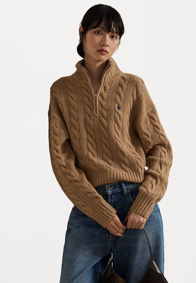 Maglione marrone a maglia cable con colletto alto, zip e polsini a coste. Abbinato a jeans in denim blu e una borsa scura.