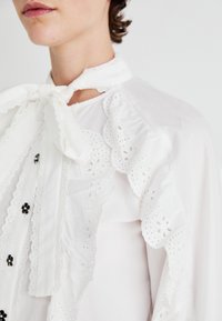 Blusa blanca con un delicado borde de encaje, lazo en el escote y acentos de flores negras en los botones delanteros. Detalles de tela texturizada visibles.