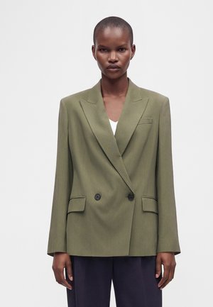 Person med kort hår iført en oversized olivengrøn dobbeltradet blazer og mørke bukser mod en ensartet lys baggrund.