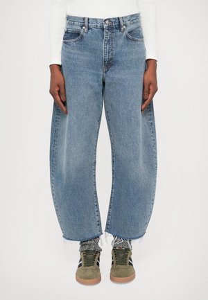 Person iført højtaljede, vide blå jeans med frynsete kanter, mønstrede sokker og grønne ruskindssneakers med sorte striber.