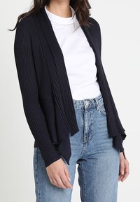 Cardigan côtelé bleu marine à ouverture sur le devant, avec un design fluide et des bords drapés, porté sur un haut blanc avec un jean bleu taille haute.