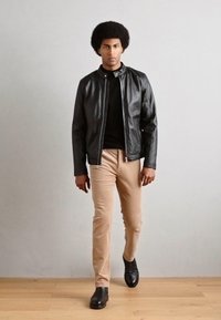 Jachetă din piele neagră, cămașă neagră ajustată, pantaloni bej și pantofi închiși. Modelul stă pe o podea din lemn, pe un fundal simplu.