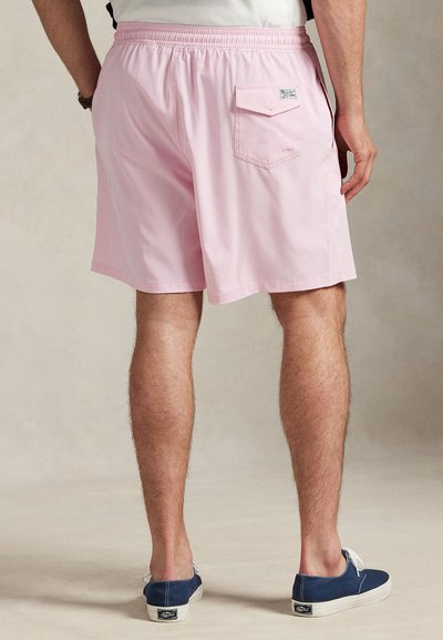 Bărbat purtând pantaloni scurți roz deschis cu buzunar pe spate și adidași casual bleumarin, stând pe un fundal neutru.