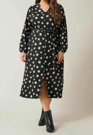 Frau trägt ein schwarzes knielanges Kleid mit beigen Polka-Dots, langen Ärmeln, Taillenschleife, vorderem Schlitz und schwarzen Stiefeletten.