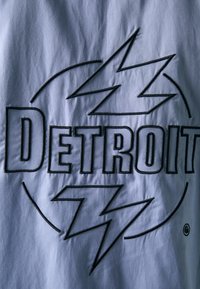 Harmaa kangas, jossa on kohopainettuja mustia kirjaimia muodostaen sanan "DETROIT" ja salamanmuotoisia koristeita. Kangas näyttää sileältä ja hieman teksturoitulta.