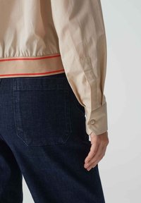 Crèmekleurig shirt met een getailleerde zoom en een rode accentband. Donkerblauwe denim broek met een achterzak, waarop een hand op de heup rust.