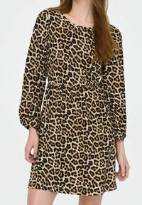Frau in einem langärmeligen Leopardenmuster-Kleid mit tailliertem Schnitt, die vor einem schlichten weißen Hintergrund steht.