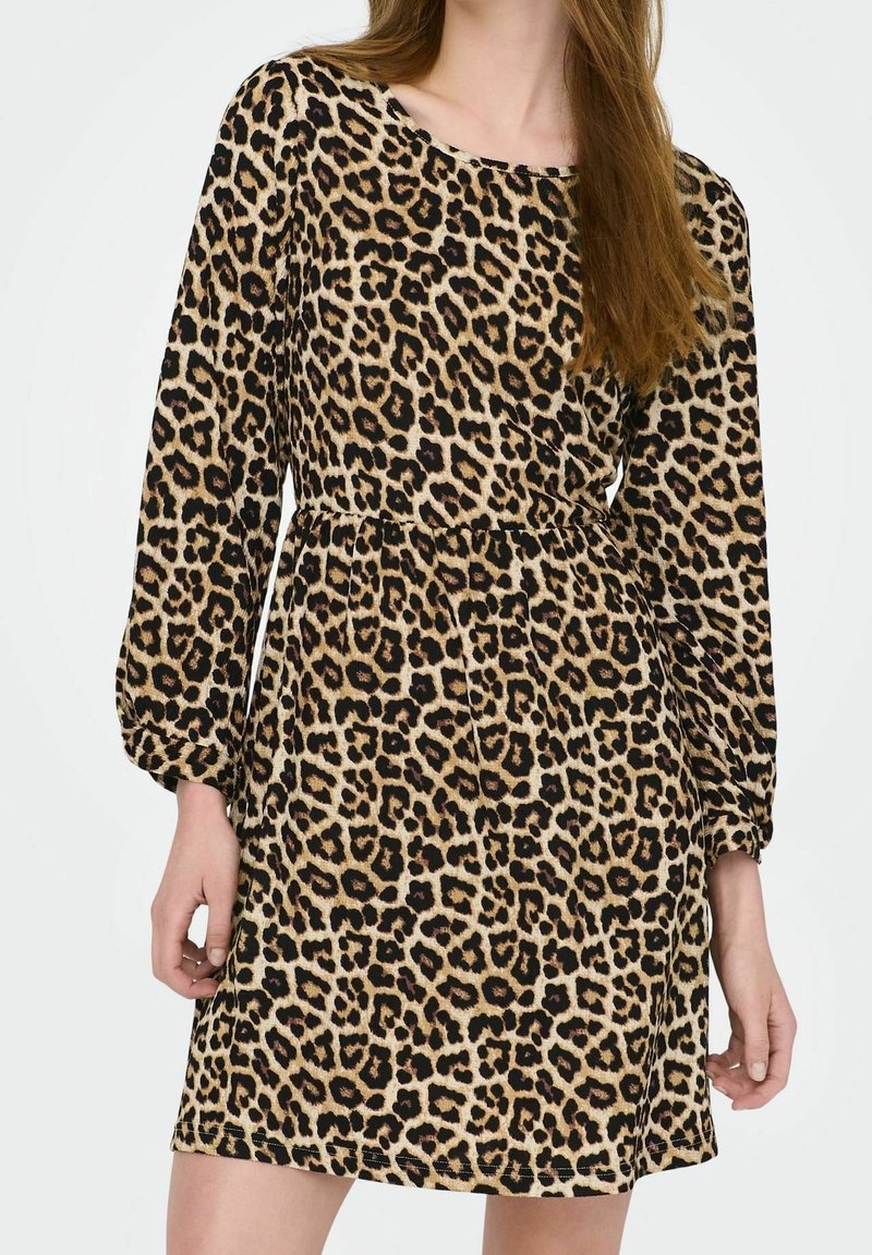 Frau in einem langärmeligen Leopardenmuster-Kleid mit tailliertem Schnitt, die vor einem schlichten weißen Hintergrund steht.