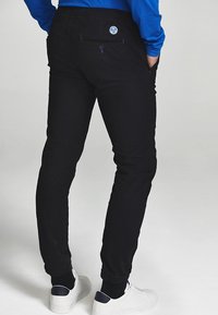 Czarne, dopasowane spodnie o gładkiej fakturze, z tylną kieszenią i okrągłym logo. W połączeniu z białymi butami.