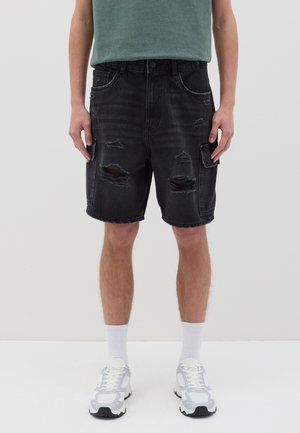Schwarze Denim-Shorts mit Distress-Details und mehreren Taschen, kombiniert mit einem grünen T-Shirt und weißen Sportschuhen sowie Socken.