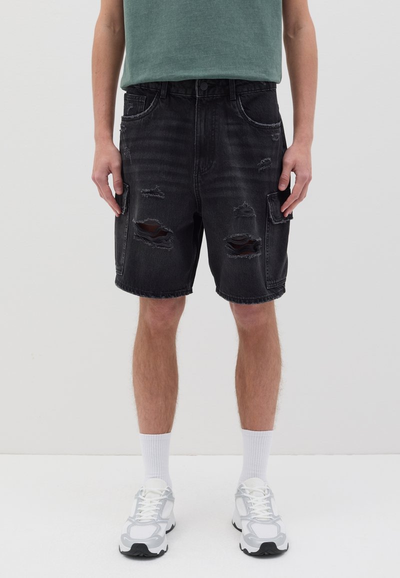 Shorts en denim noir avec des détails usés et plusieurs poches, associés à un T-shirt vert et des chaussures de sport blanches avec des chaussettes.