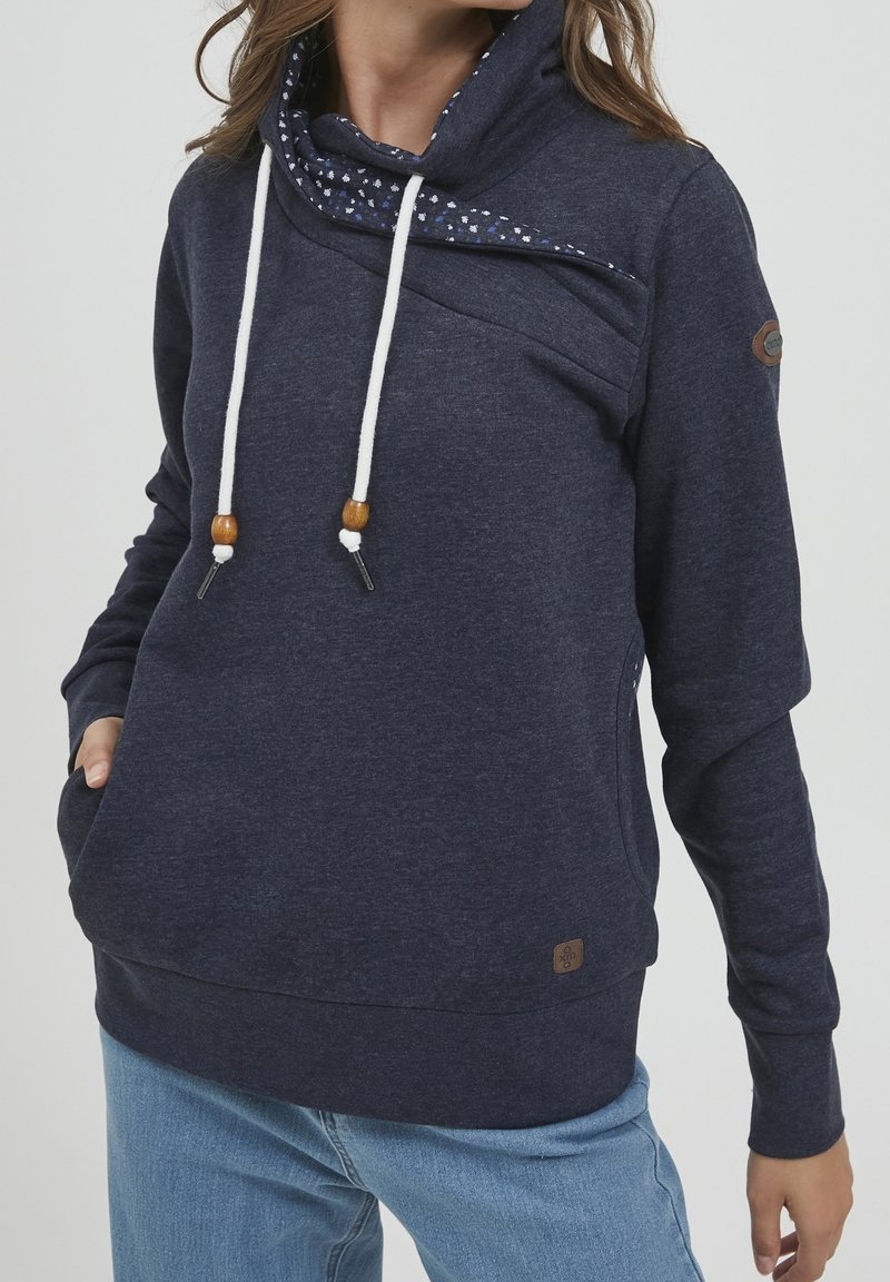 Femme portant un sweat à capuche bleu marine avec des cordons blancs et un col intérieur à fleurs, associée à un jean bleu clair, main dans la poche.