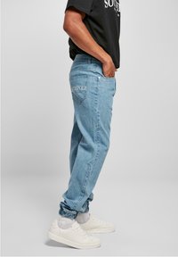 Jeans de denim azul claro con un corte relajado, que cuentan con bolsillos laterales y "SOUTHPOL" bordado en el bolsillo trasero. Combinados con zapatillas blancas.