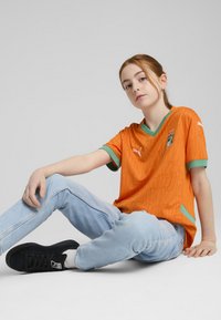 Oranžový krátký rukávový dres s zelenými akcenty a strukturovaným vzorem, kombinovaný se světlými džíny a černými teniskami.