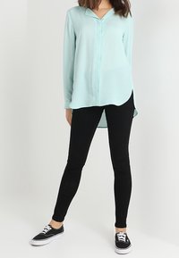 Blusa de manga larga en azul claro, con cuello, botonera y dobladillo curvado, combinada con jeans negros ajustados y zapatillas negras.