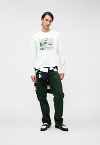 Camicia bianca a maniche lunghe con grafica del logo verde Lacoste, abbinata a pantaloni verde scuro e un maglione a righe navy/bianco legato in vita. Scarpe nere.