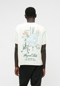 Witte katoenen T-shirt met een grafisch ontwerp van een blauwe stoel en tafel met bloemenprint op de achterkant. Inclusief naam en adres van de studio.