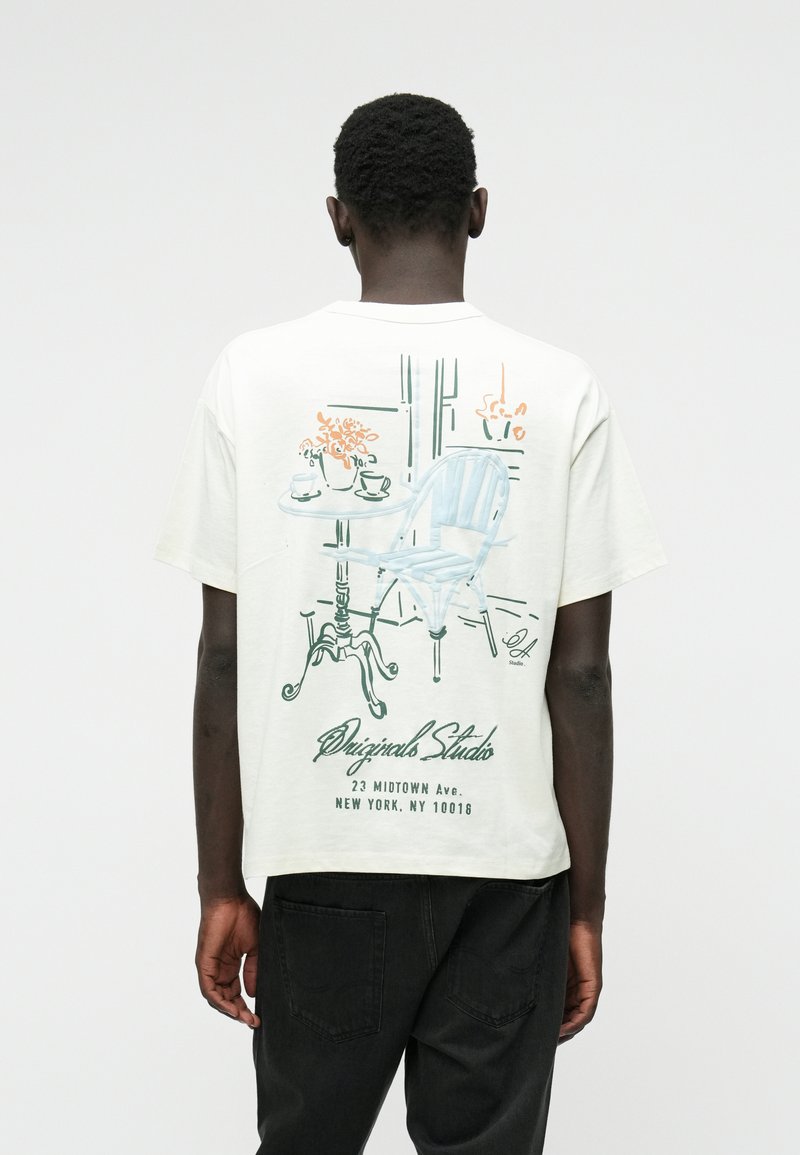 Witte katoenen T-shirt met een grafisch ontwerp van een blauwe stoel en tafel met bloemenprint op de achterkant. Inclusief naam en adres van de studio.