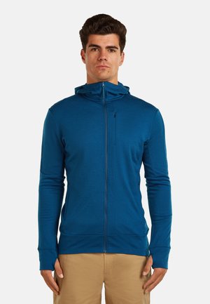 Man draagt een blauwe hoodie met rits en duimgaten, gecombineerd met beige broek, staand tegen een effen witte achtergrond.