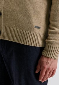 Gehaakte cardigan in beige met een ribbelzoom en boorden; voorzien van zwarte knopen en een klein metalen logo label nabij de onderkant.