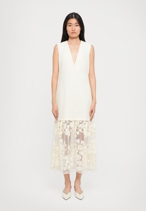 Mouwloze jurk met een diepe V-hals van off-white linnen, gelaagd met een sheer, geborduurde bloemen tulle rok. Wit, puntige schoenen.