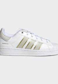 Bílé tenisky se zlatými akcenty, se třemi pruhovanými vzory, texturovanou kůží a gumovou podešví. Na boku je označeny jako "SUPERSTAR".