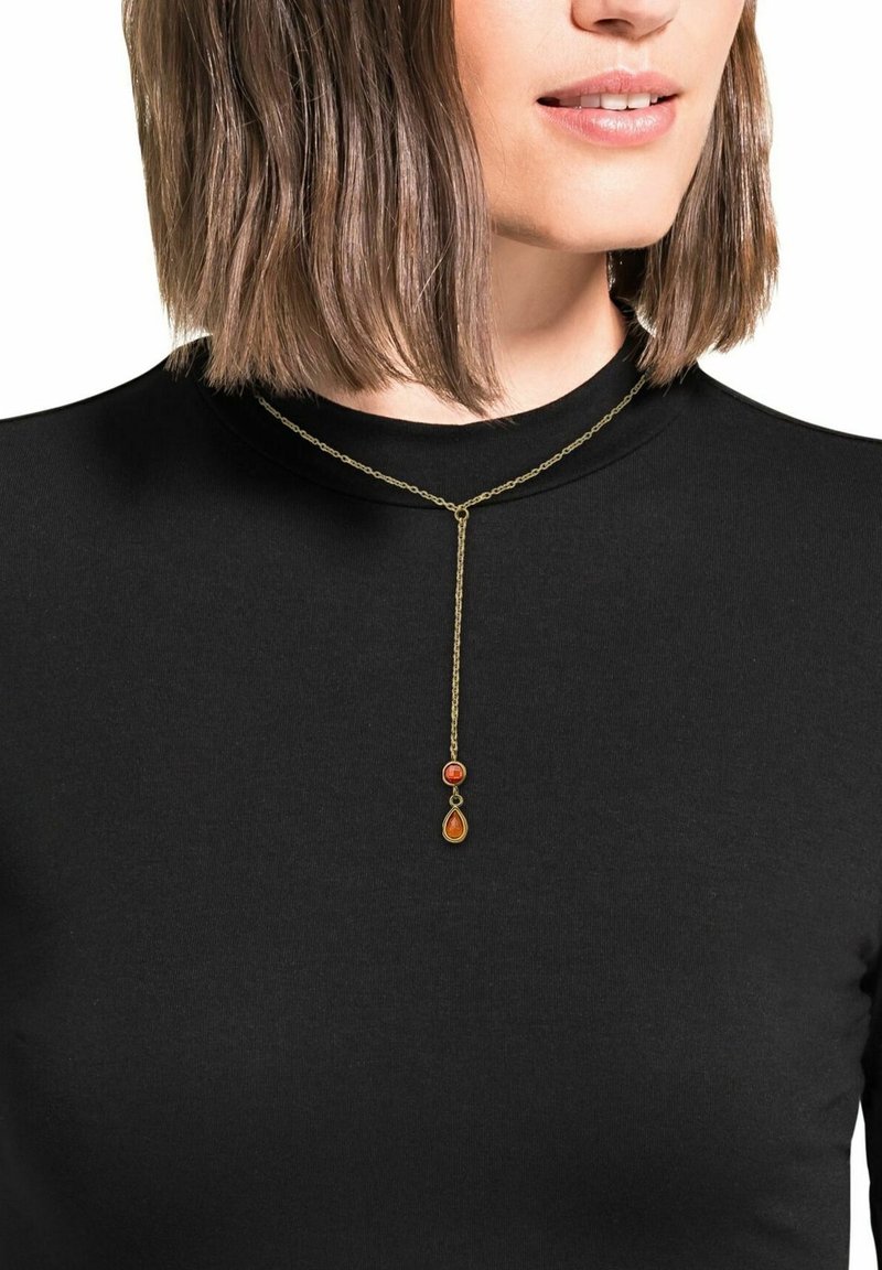 Collana in oro con due pendenti, uno arancione e uno rosso, di forma a goccia con un accento circolare, indossata sopra una maglietta nera.
