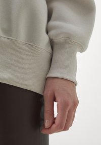 Light gray sweatshirt with ribbed cuffs and hem, detailed stitching, worn over brown leather pants. Hand resting on thigh.