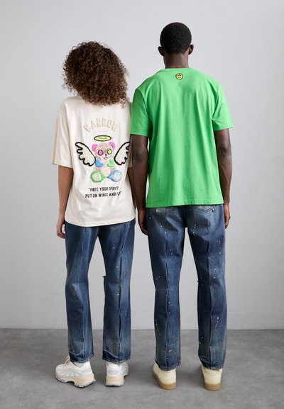 T-shirt crème surdimensionné avec un graphique coloré de ours et le texte "LIBÉREZ VOTRE ESPRIT" ; T-shirt vert ; les deux associés avec un jean bleu déchiré et des baskets.