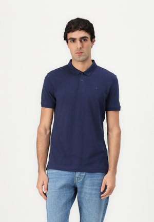 Polo shirt bleu marine en mélange de coton, avec un col classique, trois boutons et un logo discret sur la poitrine. Associé à un jean bleu clair.