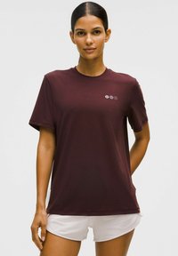 Egy maroon színű pólót viselő nő, melyen kis logó található a mellrészen, és fehér sportnadrágot hord, egy sima világos háttér előtt áll.