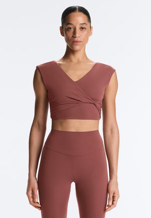 Frau trägt ein ärmelloses, rostfarbenes Wickel-Crop-Top und passende, hoch taillierte Leggings, steht vor einem schlichten hellen Hintergrund.