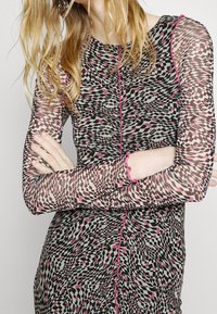 Pitkähihaista mekkoa, jossa on läpinäkyvää kangasta ja geometrinen kuvio mustasta, vaaleanpunaisesta ja beigeestä, ja reunat on koristeltu vaaleanpunaisella aallotuksella.