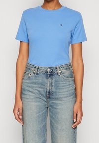  person iført en tætsiddende lys blå kortærmet skjorte, stoppet ned i højtaljede, lysvaskede blå jeans med synlige knapper og lommer.