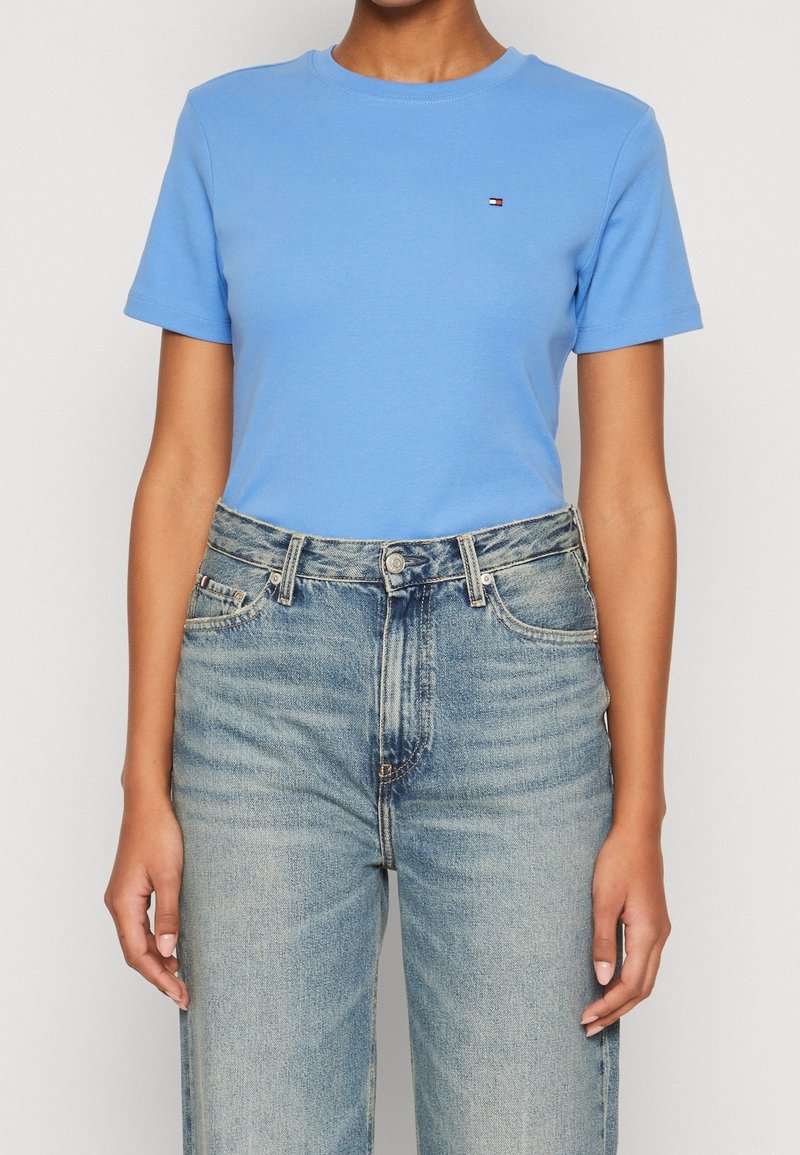 person iført en tætsiddende lys blå kortærmet skjorte, stoppet ned i højtaljede, lysvaskede blå jeans med synlige knapper og lommer.