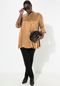 Blusa di raso color caramello con colletto a camicia e maniche a campana, abbinata a pantaloni neri attillati e una borsa a tracolla nera.