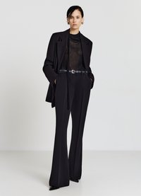 Costume noir sur mesure comprenant un blazer long et ouvert et un pantalon évasé taille haute. Top noir transparent en dessous, complété par une ceinture noire.