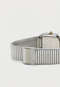 Ασημένιο μεταλλικό ρολόι με ορθογώνιο καντράν, εύκαμπτο μπρασελέ με κρίκους, κούμπωμα με λογότυπο κροκόδειλου και χρυσόχρωμο κουρδιστήρι.