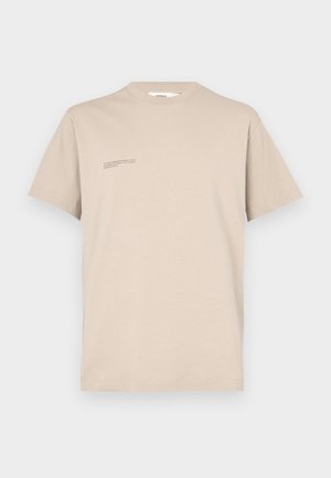 Lyhythihainen beige t-paita, joka on valmistettu luomupuuvillasta. Oikealla rintapielessä on pieni painettu tekstisuunnittelu. Pehmeä pinta.